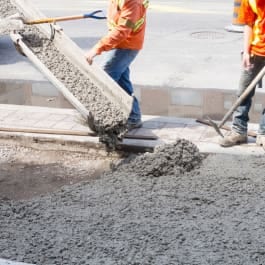 Workers pouring concrete onto a sidewalk from a chute, with tools nearby.