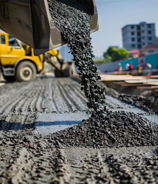 Close-up of a bulldozer pouring wet concrete onto a construction site, with tire tracks visible. A building and a yellow truck are in the background.