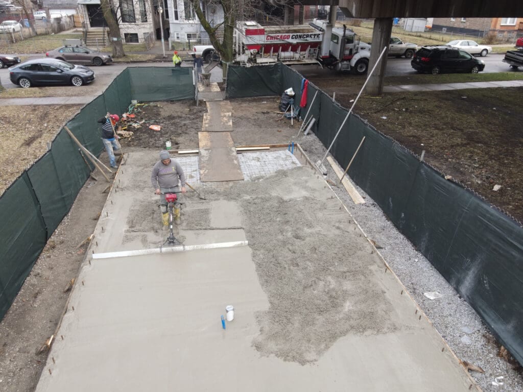 Construction workers pour and smooth concrete on a rectangular foundation surrounded by a green fence, with vehicles and trees in the background.