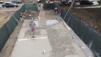 Construction workers pour and smooth concrete on a rectangular foundation surrounded by a green fence, with vehicles and trees in the background.