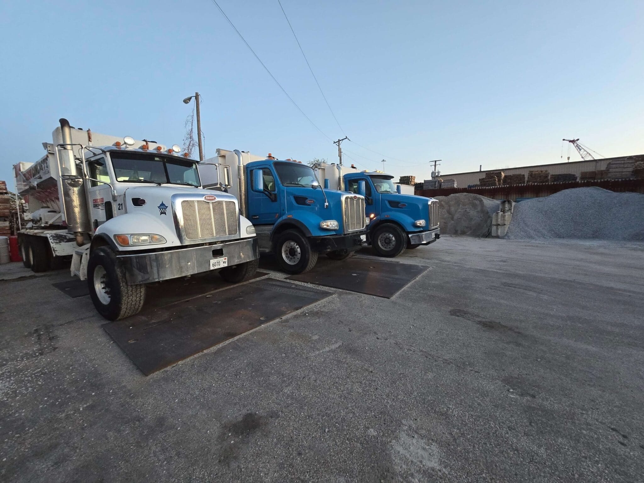 Three parked trucks on an industrial lot with piles of gravel in the background. The sky is clear at dusk.