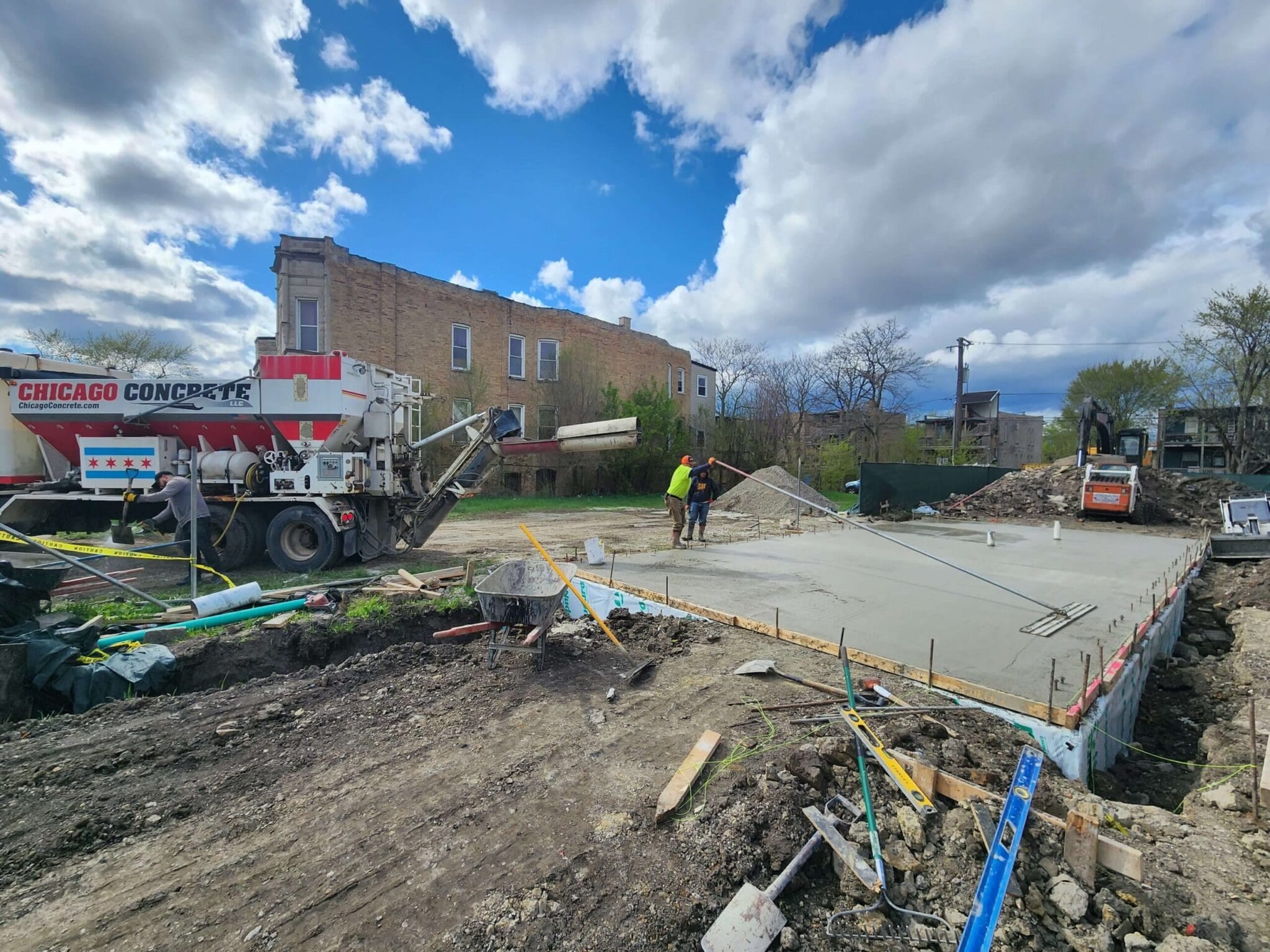 Construction workers are leveling freshly poured concrete on a foundation at a building site. A concrete truck is parked nearby under a cloudy sky.