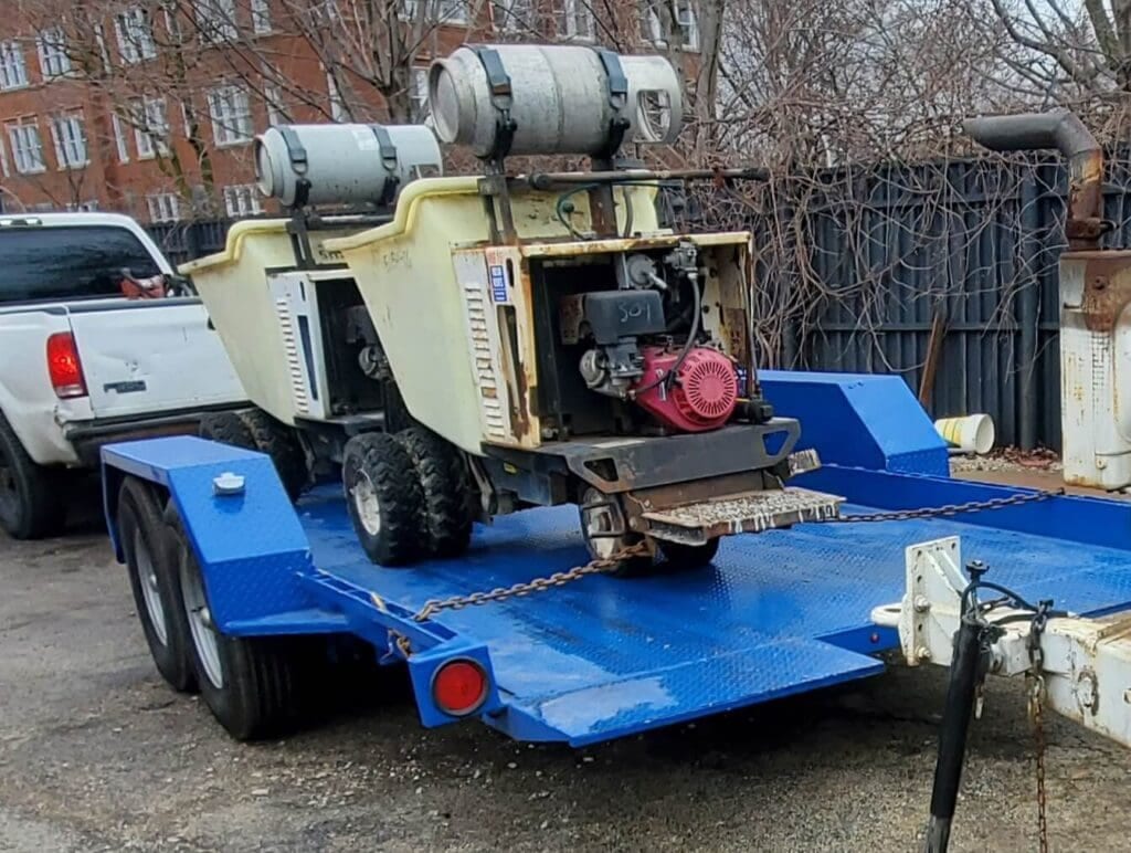 Two utility carts with gas tanks are secured on a blue trailer, attached to a white truck on a paved area near a fence.