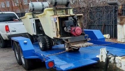 Two utility carts with gas tanks are secured on a blue trailer, attached to a white truck on a paved area near a fence.