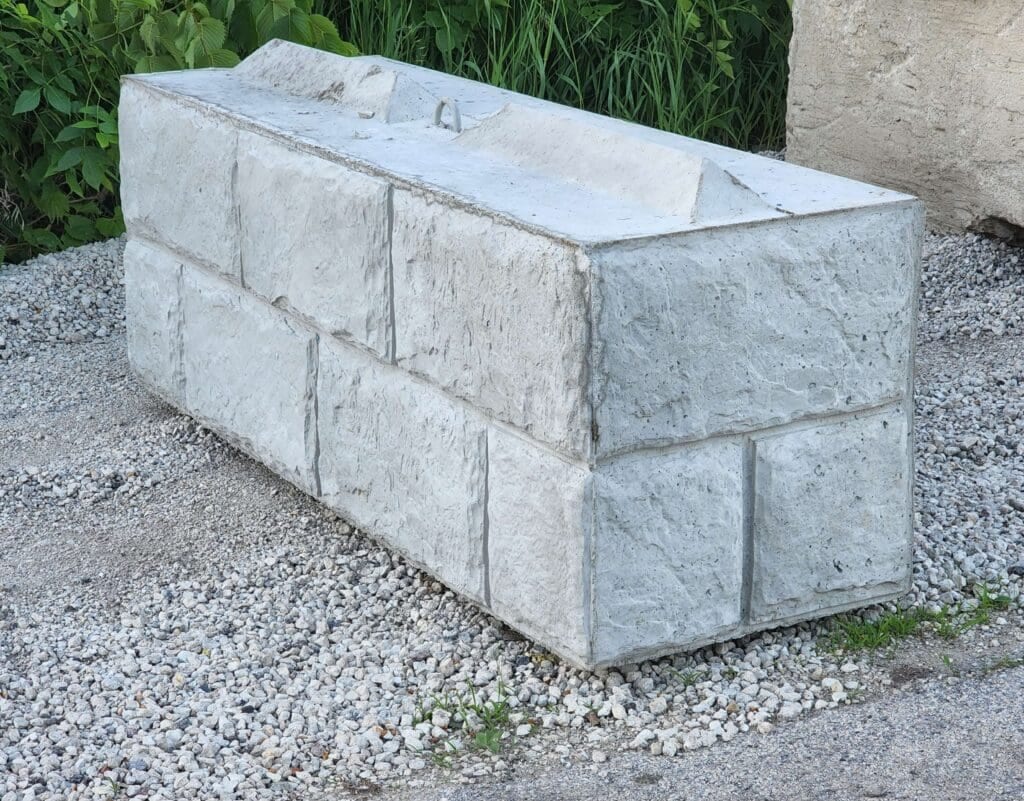 Concrete block with a rectangular shape, resting on gravel, surrounded by greenery and partially another structure.