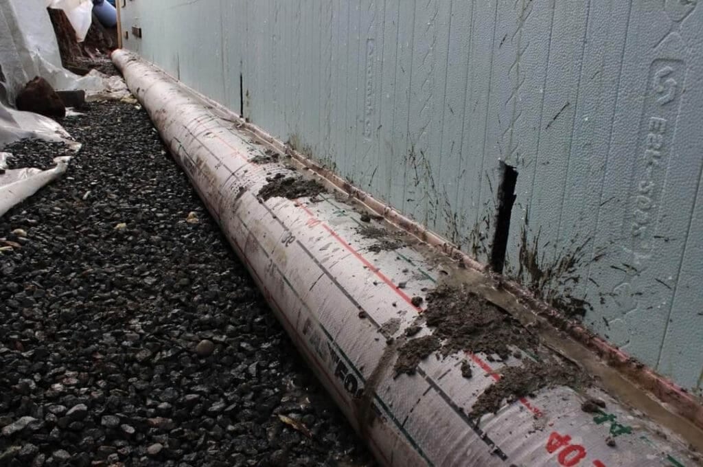 A large cylindrical pipe covered in a white protective film is laid next to a light blue insulated wall. Gravel is spread around the pipe, and a small cut is visible in the wall.