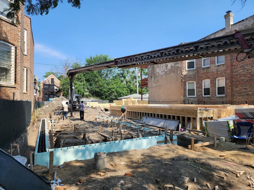 Construction site with workers operating machinery and laying foundation surrounded by brick buildings and trees under clear blue sky.