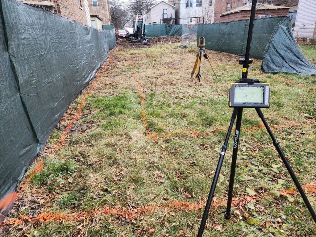 A grassy area marked with orange lines, enclosed by green fencing. Two surveying tripods with equipment are set up in the space.