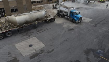 A blue truck is parked next to a tanker trailer in an industrial lot, with buildings and machinery in the background. Workers and vehicles are visible in the distance.