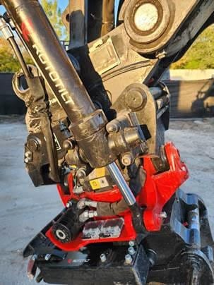 Close-up of industrial machinery components, featuring a black and red Rototilt attachment with hydraulic connections and bolts, set against a blurred outdoor background.