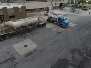 A blue truck is parked next to a tanker trailer in an industrial lot, with buildings and machinery in the background. Workers and vehicles are visible in the distance.